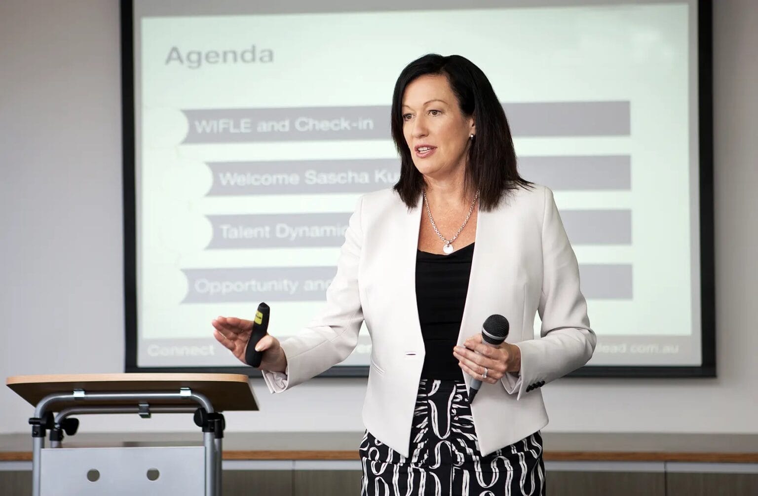 Margo Halbert holding a microphone, making a presentation in front of a projection screen.