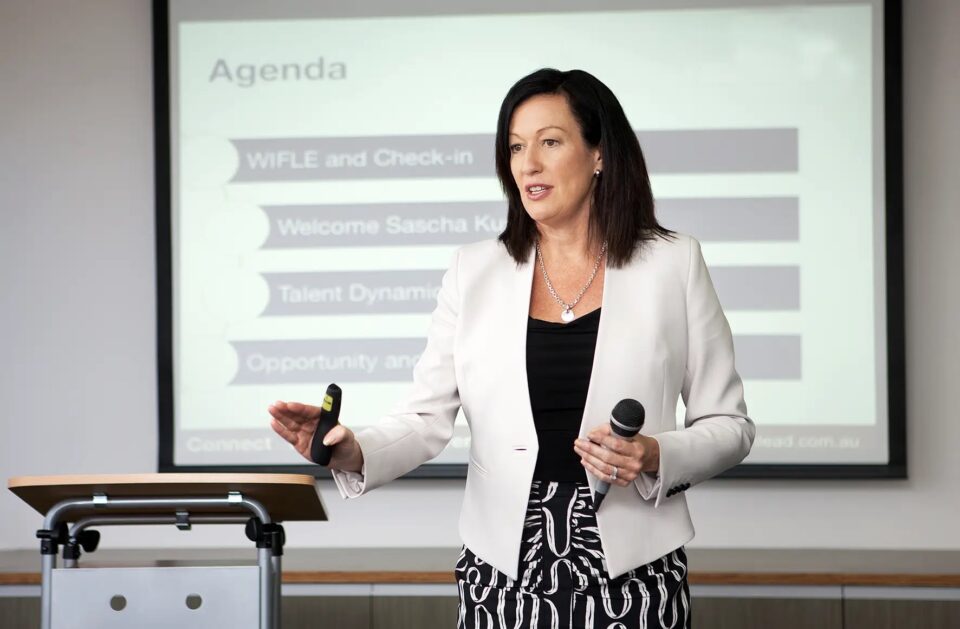 Margo Halbert holding a microphone, making a presentation in front of a projection screen.